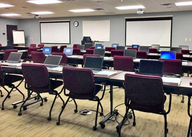 Photo of the training room with laptops on three rows of desks facing a podium and projector screens